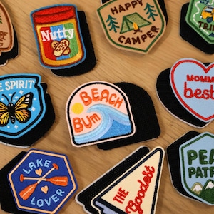 May include: A collection of embroidered patches with various designs and text. Patches include a jar of peanut butter labeled "Nutty", a camping scene with "Happy Camper", and a beach scene with "Beach Bum". Other patches feature a butterfly, paddles, and mountains.