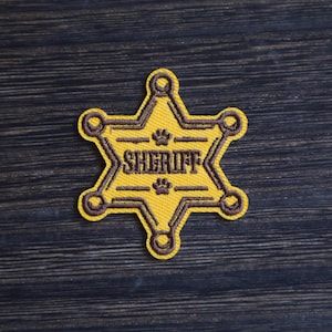 May include: A yellow sheriff badge patch with brown embroidered details. The badge is star-shaped with rounded points and features the word "SHERIFF" in capital letters. Paw prints are above and below the word.