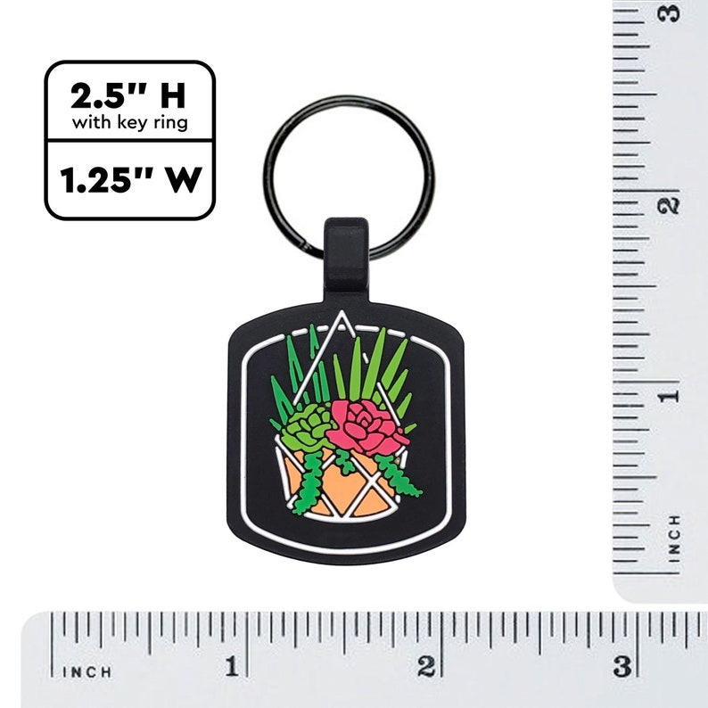 May include: Black keychain with a succulent plant design. The keychain is 2.5 inches high with the key ring and 1.25 inches wide. The design features a hanging orange pot with green and red succulents, and green leaves.