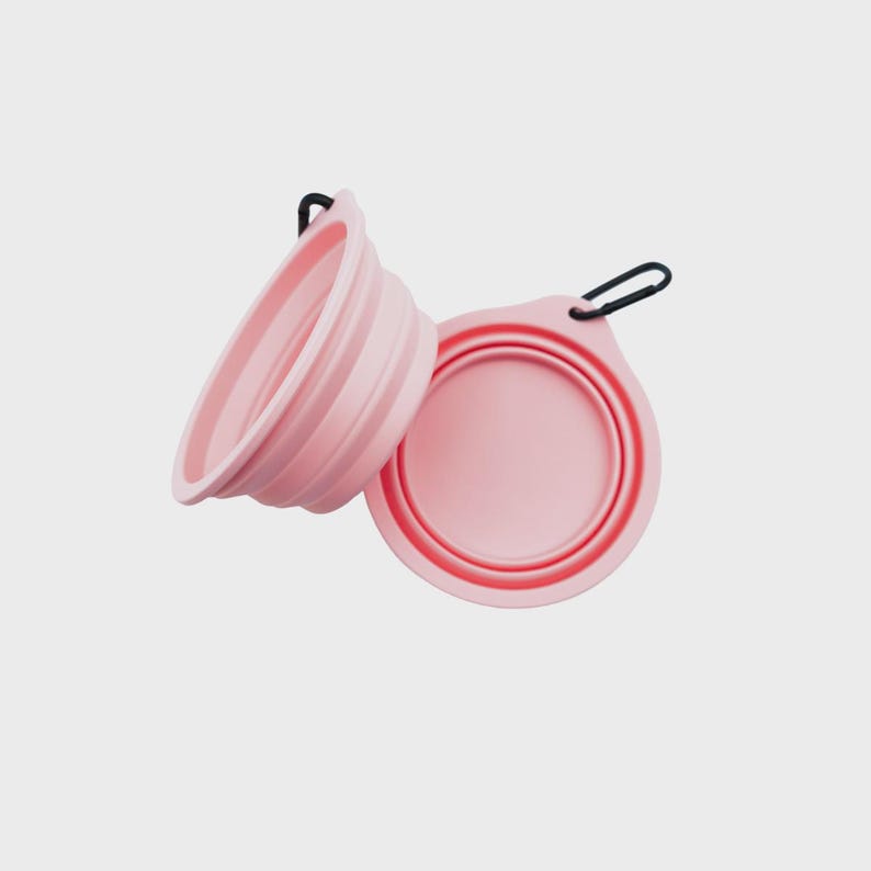 May include: Two light pink collapsible bowls made of flexible silicone. Each bowl features a circular base, ribbed sides for folding, and a small tab with a black metal carabiner attached for easy carrying and storage during travel or outdoor use.