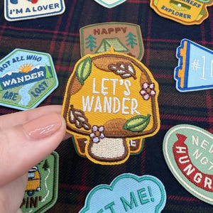 May include: A collection of embroidered patches with different designs and text. One patch features a mushroom with "LET'S WANDER", and another shows a tent with "HAPPY". Other patches say "I'M A LOVER" and "NOT ALL WHO WANDER ARE LOST".