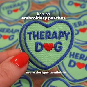 May include: A heart-shaped, light green iron-on embroidery patch with the words "THERAPY DOG" in blue, and a red heart. The patch is surrounded by a blue border. The text "iron-on embroidery patches" and "more designs available" are also visible.