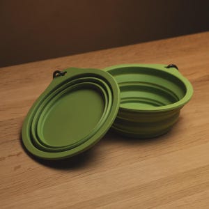 May include: A collapsible olive green silicone bowl featuring a circular base and a flexible, accordion-style pleated wall design. The bowl includes a small integrated tab with a black metal carabiner attached for convenient hanging or transport.