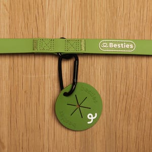 May include: A light green strap with a rectangular patch reading Besties and a black carabiner attached to a round green waste bag dispenser. The circular dispenser features a star-shaped opening, the word besties embossed, and a white logo.