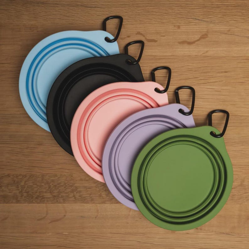May include: Five collapsible circular bowls in light blue, black, light pink, lavender, and olive green. Each bowl features a flexible, ribbed silicone construction with a flat base and a small tab at the top edge attached to a black metal carabiner.