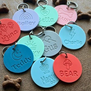 Könnte beinhalten: Eine Sammlung farbenfroher Hunde- und Katzenmarken mit verschiedenen Namen und Designs. Die runden Marken tragen Namen wie "Bailey", "Bella" und "Charlie". Jede Marke hat ein anderes Design, darunter ein Regenbogen, ein Herz, ein Knochen und ein Blitz. Sie sind an silbernen Schlüsselringen befestigt.