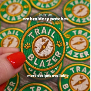 May include: Green and gold iron-on embroidery patches with the text "TRAIL BLAZER" and a compass design. The patches are round and feature paw prints. The text "iron-on embroidery patches" and "more designs available" are also visible.