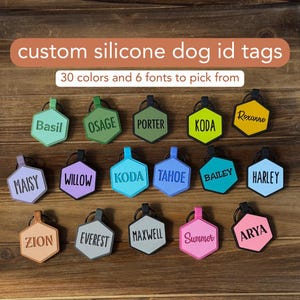 May include: A selection of colourful silicone dog ID tags with various names printed on them. The tags are shaped like hexagons and have a metal ring for attaching to a collar.