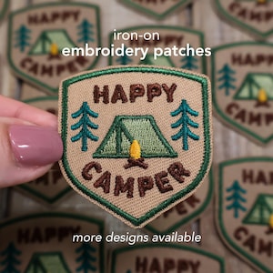 May include: Tan iron-on embroidery patch with a shield shape. The patch features the words "HAPPY CAMPER" and a green tent, two green trees, and a yellow campfire. Text on the patch reads "iron-on embroidery patches" and "more designs available."