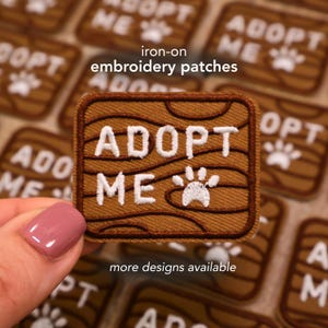 May include: Brown iron-on embroidery patch with the words "ADOPT ME" in white, along with a white paw print. The patch has a wood grain design. The text "more designs available" is also present.