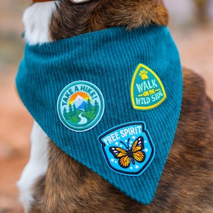 May include: A teal corduroy dog bandana with three embroidered patches. The patches read "Take a Hike!", "Walk on the Wild Side", and "Free Spirit" with a butterfly illustration. The bandana is around a dog's neck, secured by a brown leather collar.