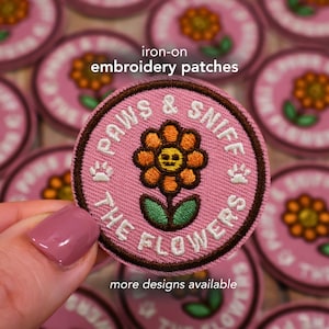 May include: Pink iron-on embroidery patch with a brown border. The patch features a smiling orange flower with a green stem and leaves. Text on the patch reads "PAWS & SNIFF THE FLOWERS". Additional text says "more designs available".