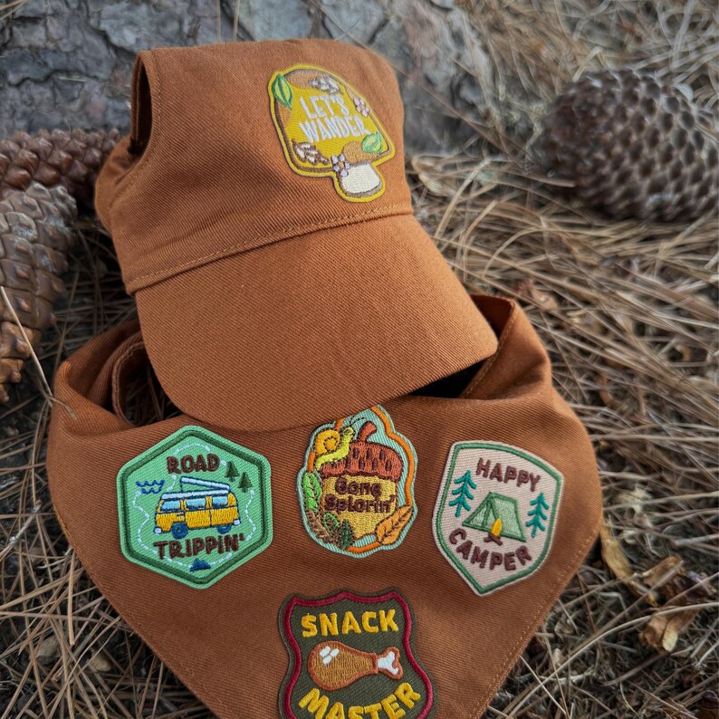 Puede incluir: Un conjunto marr&oacute;n con tem&aacute;tica de camping, compuesto por una gorra y una bandana. La gorra tiene un parche que dice "Let's Wander". La bandana tiene parches que dicen "Road Trippin'", "Gone S'lorin'", "Happy Camper" y "Snack Master".