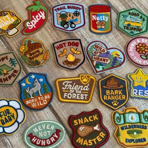 May include: A collection of embroidered patches with various designs and text. Patches include a red chili pepper, a peanut butter jar, a hot dog, and phrases like "Trail Buddy" and "Born to be Wild." The patches are in various shapes and colors.