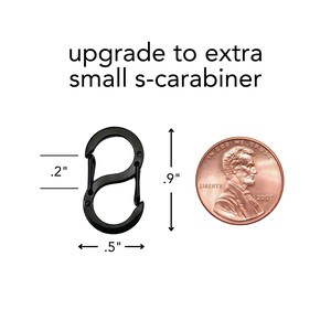 May include: Black s-carabiner, .9 inches long, .5 inches wide, and .2 inches thick. The text "upgrade to extra small s-carabiner" is above the carabiner. A penny is next to the carabiner for size comparison.