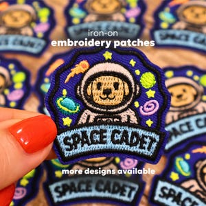 May include: Embroidered iron-on patch featuring a cartoon dog astronaut in a space suit. The patch is blue with yellow stars, a planet, and the words "SPACE CADET". The text "iron-on embroidery patches" is also visible.