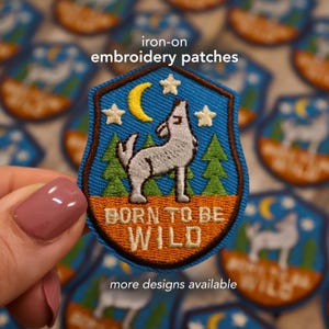 May include: Iron-on embroidery patch with a wolf howling at a yellow crescent moon and stars. The patch has a blue background with green trees and the text "BORN TO BE WILD" in orange.