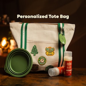 May include: A beige canvas tote bag with green striped handles, featuring embroidered patches of a tree, a wilderness explorer badge, and a trail blazer emblem. A green silicone bowl and two tubes of Besties Butter are placed in front.