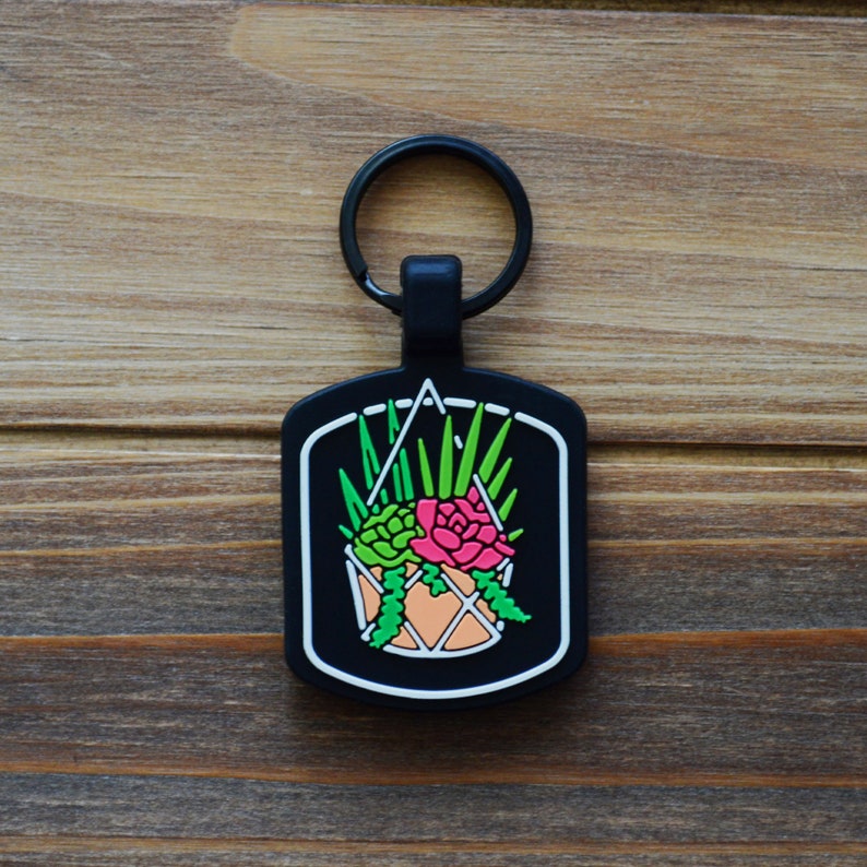 May include: Black rectangular keychain featuring a colorful succulent design. The design includes green, pink, and orange succulents in a hanging pot, outlined in white against a black background. The keychain has a black ring.