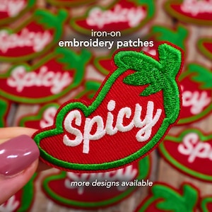 May include: Red and green iron-on embroidery patches in the shape of a chili pepper. The word "Spicy" is embroidered in white on the pepper. The text "iron-on embroidery patches" and "more designs available" are also visible.