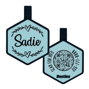 May include: Two hexagonal blue and black keychains with a black and white QR code on one and the name "Sadie" with a heart and floral design on the other. The keychain with the QR code also has the text "Scan for Info" and "Besties" on it. The text on the keychain with the QR code is "555-777-8888".