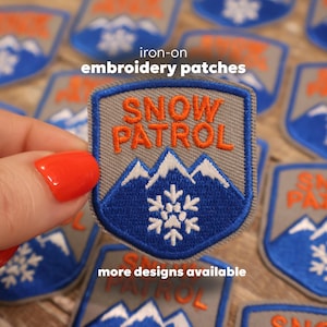 May include: Iron-on embroidery patches with a shield design. The patches feature the words "SNOW PATROL" in orange, a blue mountain range, and a white snowflake. The patches are gray with blue and white accents.