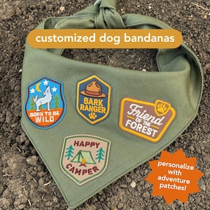 May include: Olive green dog bandana with four embroidered patches. Patches include a howling wolf, a ranger hat, a paw print, and a tent. Text on the patches reads "Born to be Wild", "Bark Ranger", "Friend of the Forest", and "Happy Camper".