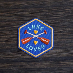 May include: A blue, hexagon-shaped patch with the words "LAKE LOVER" in yellow. Two crossed red and white oars are in the center, with two yellow hearts. The patch has a yellow border and is on a dark wood surface.