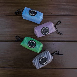 May include: Four dog waste bag dispensers in different colors: light blue, purple, green, and gray. Each dispenser has a black clip and a round black label with the word "besties" printed on it.