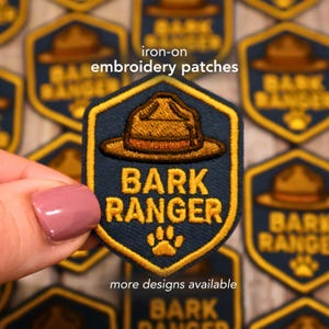 May include: A navy blue and gold iron-on embroidery patch with the text "BARK RANGER" and a paw print. The patch features a ranger hat design. The text "iron-on embroidery patches" and "more designs available" are also visible.