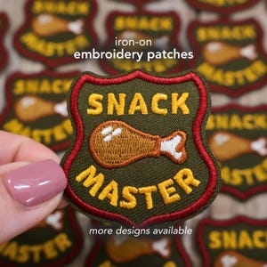 May include: An iron-on embroidery patch with a shield design. The patch features the words "SNACK MASTER" in yellow, with a cartoon drumstick in the center. The patch is green with a red border. The text "more designs available" is at the bottom.