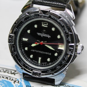 Puede incluir: Reloj de pulsera negro con bisel plateado y correa de cuero negro. La esfera negra tiene números blancos, agujas luminosas y el texto "АВТОПОДЗАВОД". También tiene una escala taquimétrica.