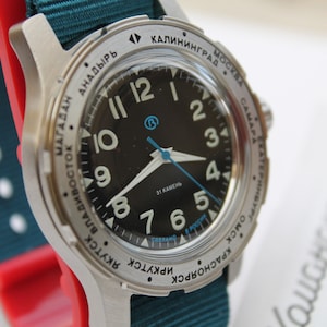 Op de afbeelding: Een zilveren polshorloge met een zwarte wijzerplaat en witte cijfers. Het horloge heeft een draaibare bezel met stadsnamen die rond de rand zijn gedrukt. Het horloge zit op een tealgroene nylon band.