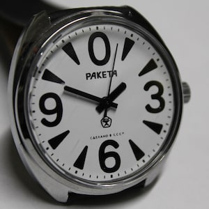 Puede incluir: Un reloj de pulsera plateado con una esfera blanca y números negros. La esfera del reloj dice "ПАКЕТА" y "СДЕЛАНО В СССР".
