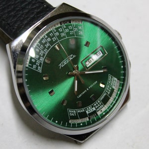 Puede incluir: Un reloj de pulsera analógico verde con función de calendario. La esfera del reloj tiene un bisel y agujas plateadas. La esfera del reloj también tiene una pantalla del día de la semana y una pantalla de fecha. La correa del reloj es de cuero negro.