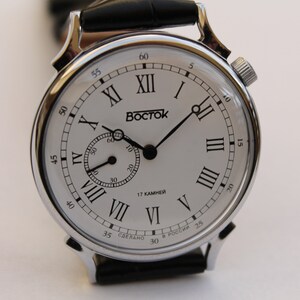 Puede incluir: Un reloj de pulsera plateado con una esfera blanca y marcadores de hora en n&uacute;meros romanos negros. El reloj tiene una correa de cuero negra y un peque&ntilde;o dial de segundero. La esfera del reloj tambi&eacute;n incluye el texto "BOCTCK" y "17 KAMHEЙ".