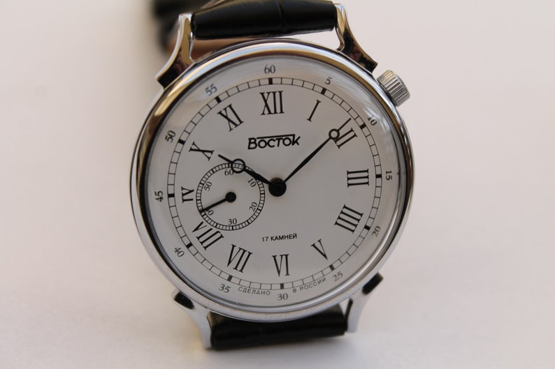 Puede incluir: Un reloj de pulsera plateado con una esfera blanca y marcadores de hora en n&uacute;meros romanos negros. El reloj tiene una correa de cuero negra y un peque&ntilde;o dial de segundero. La esfera del reloj tambi&eacute;n incluye el texto "BOCTCK" y "17 KAMHEЙ".