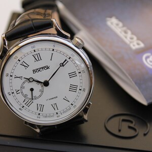 Puede incluir: Un reloj de pulsera plateado con una esfera blanca y marcadores de hora en n&uacute;meros romanos negros. El reloj tiene una correa de cuero negra y un peque&ntilde;o dial de segundero. La esfera del reloj tambi&eacute;n incluye el texto "Восток" y "17 камней".