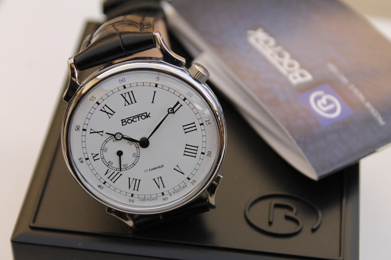 Puede incluir: Un reloj de pulsera plateado con una esfera blanca y marcadores de hora en n&uacute;meros romanos negros. El reloj tiene una correa de cuero negra y un peque&ntilde;o dial de segundero. La esfera del reloj tambi&eacute;n incluye el texto "Восток" y "17 камней".