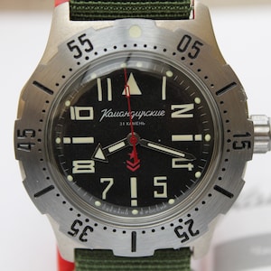 Può includere: Un orologio da polso nero e argento con un cinturino verde. Il quadrante dell'orologio ha numeri bianchi e una lancetta dei secondi rossa. Il quadrante dell'orologio ha anche il testo "Komandirskie 31 камень".