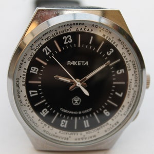 Könnte beinhalten: Eine silberne Armbanduhr mit schwarzem Zifferblatt und weißen Markierungen. Das Zifferblatt der Uhr verfügt über eine Weltzeitanzeige mit Städtenamen am Rand. Das Zifferblatt der Uhr enthält auch die Wörter "RAKETA" und "СДЕЛАНО В СССР".