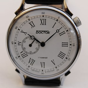 Puede incluir: Un reloj de pulsera plateado con una esfera blanca y marcadores de hora en n&uacute;meros romanos negros. El reloj tiene un peque&ntilde;o dial de segundero en la posici&oacute;n de las 3 en punto. La esfera del reloj tambi&eacute;n tiene el texto "Восток" y "17 КАМНЕЙ".