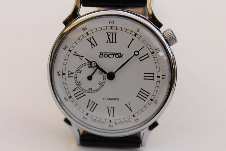 Puede incluir: Un reloj de pulsera plateado con una esfera blanca y marcadores de hora en n&uacute;meros romanos negros. El reloj tiene un peque&ntilde;o dial de segundero en la posici&oacute;n de las 3 en punto. La esfera del reloj tambi&eacute;n tiene el texto "Восток" y "17 КАМНЕЙ".