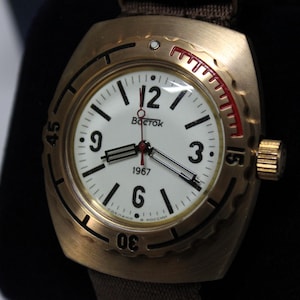 May include: A gold-toned wristwatch with a white face, black numerals, and black hands. The watch face displays the brand name "BOCTOK" and the year "1967". The watch has a brown strap and a red accent on the bezel.