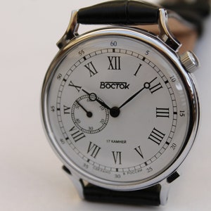 Puede incluir: Un reloj de pulsera plateado con una esfera blanca y marcadores de hora romanos negros. El reloj tiene una correa de cuero negra y un peque&ntilde;o dial de segundero. La esfera del reloj tambi&eacute;n incluye el texto "Восток" y "17 КАМНЕЙ".