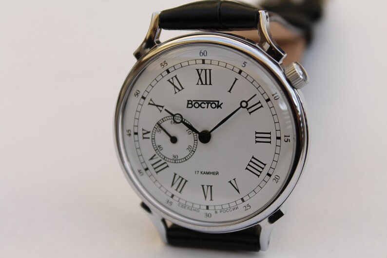 Puede incluir: Un reloj de pulsera plateado con una esfera blanca y marcadores de hora romanos negros. El reloj tiene una correa de cuero negra y un peque&ntilde;o dial de segundero. La esfera del reloj tambi&eacute;n incluye el texto "Восток" y "17 КАМНЕЙ".