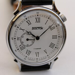 Puede incluir: Un reloj de pulsera plateado con una esfera blanca y marcadores de hora en n&uacute;meros romanos negros. El reloj tiene una correa de cuero negro y un peque&ntilde;o dial de segundero. La esfera del reloj tambi&eacute;n incluye el texto "17 KAMHEЙ" y "СДЕЛАНО В РОССИИ".