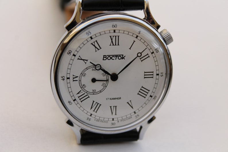 Puede incluir: Un reloj de pulsera plateado con una esfera blanca y marcadores de hora en n&uacute;meros romanos negros. El reloj tiene una correa de cuero negro y un peque&ntilde;o dial de segundero. La esfera del reloj tambi&eacute;n incluye el texto "17 KAMHEЙ" y "СДЕЛАНО В РОССИИ".