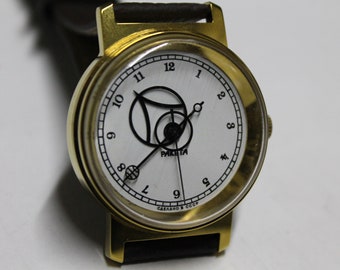 May include: A gold-tone wristwatch with a white face and black hands. The watch face features a unique design with a circle and a line. The watch face also includes the text "РАКЕТА" and "СДЕЛАНО В СССР".