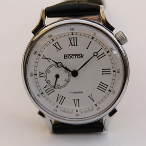 Puede incluir: Un reloj de pulsera plateado con una esfera blanca y marcadores de hora en n&uacute;meros romanos negros. El reloj tiene una correa de cuero negra y un peque&ntilde;o dial de segundero. La esfera del reloj tambi&eacute;n incluye el texto "BOCTOK" y "17 КАМНЕЙ".